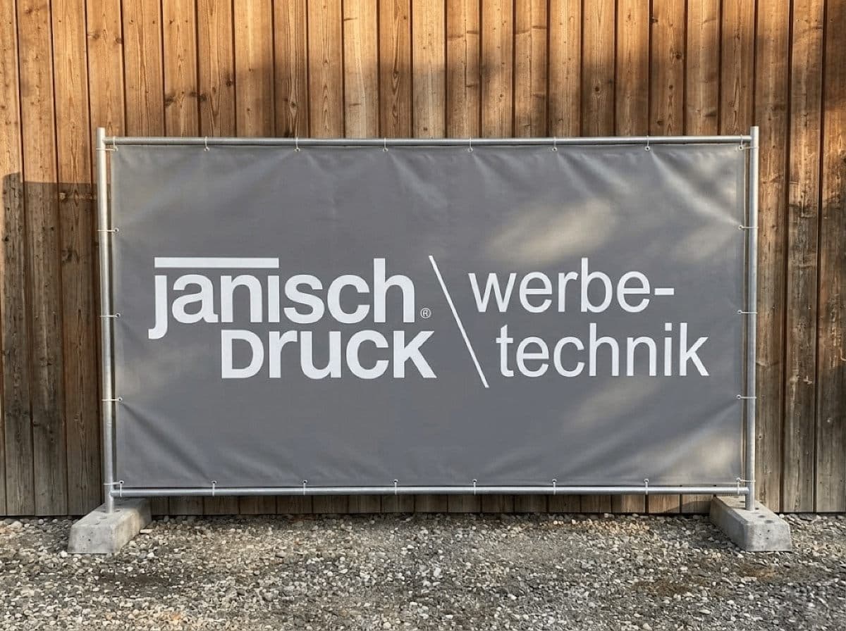 Bauzaunbanner mit Janisch-Werbetechnik-Logo an einem Metallzaun vor einer Holzfassade.